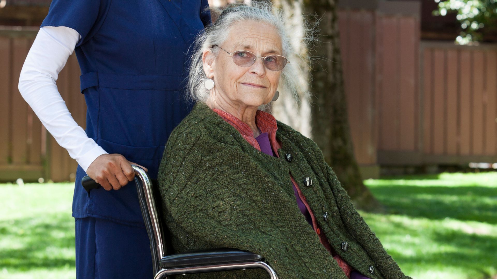 mujer mayor en silla de ruedas mirando a cámara y paseando con una cuidadora en un jardín