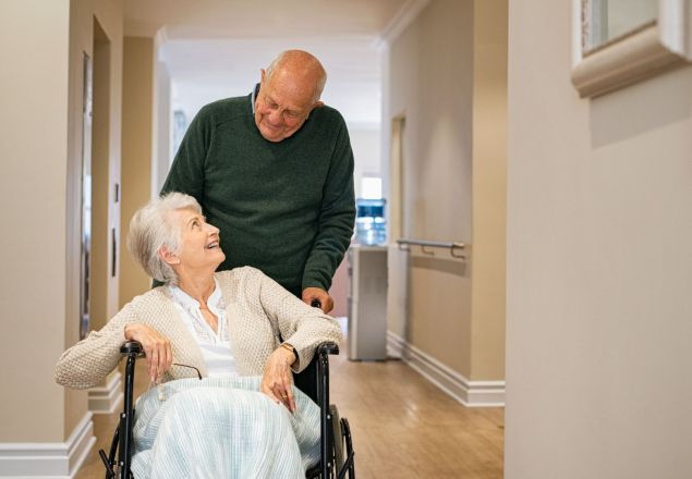 Hombre mayor acompañando a una mujer mayor en silla de ruedas por el pasillo de una residencia